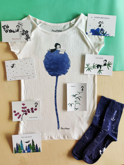 Ein Geschenk-Paket bestehend aus einem weißem Bio-Baumwolle T-Shirt mit Blumenbuch-Print (einem lesenden Mädchen, das auf einer blauen Blume liegt), 7 unterschiedlichen Grußkarten und einem Paar blaue Socken mit weißen Fischen und Ankermotiv.