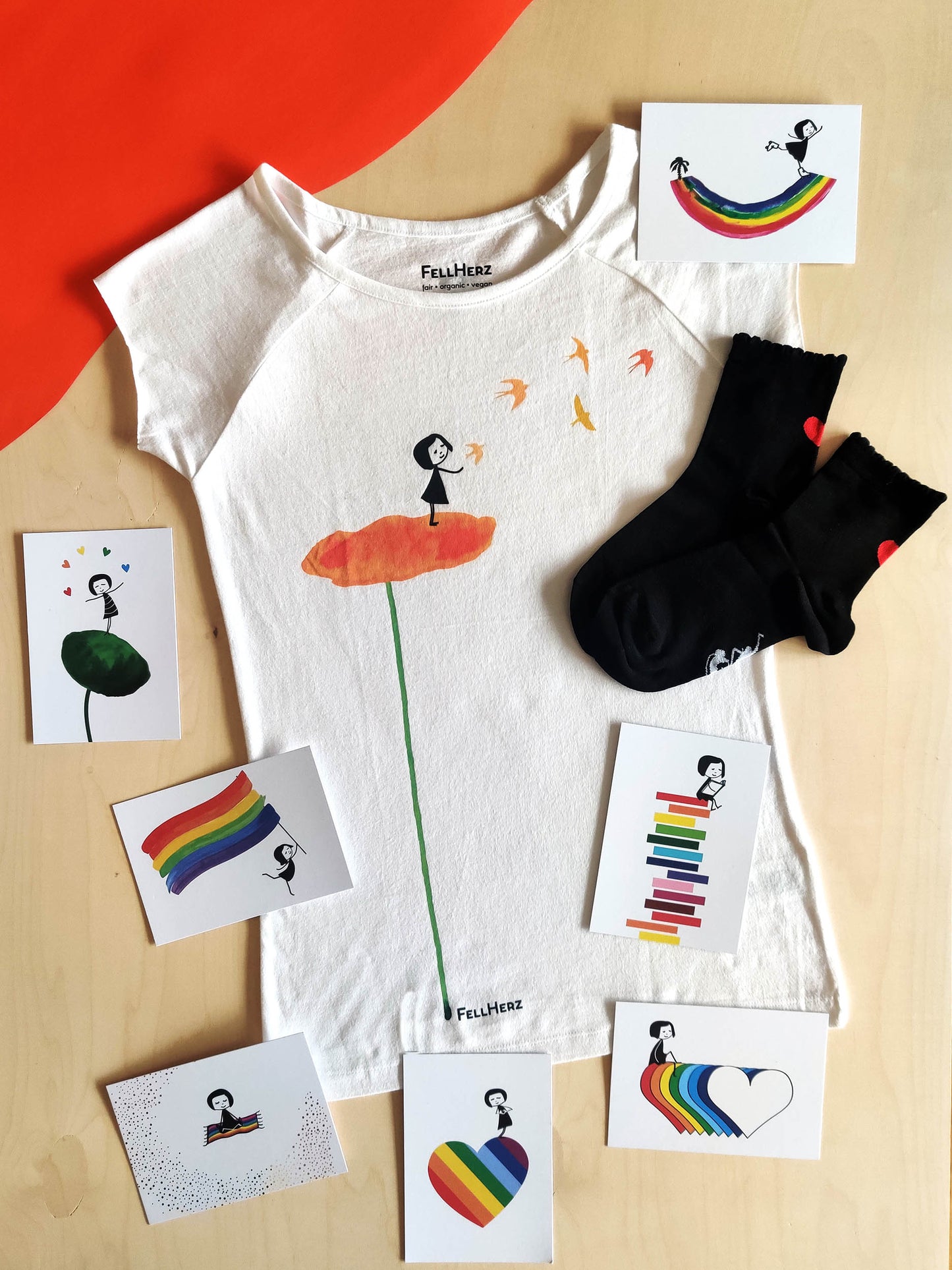 Ein Geschenk-Paket bestehend aus einem weißem Bio-Baumwolle T-Shirt mit orangem Blumenmotiv, auf der Blume steht ein Mädchen und Vögel fliegen, 7 unterschiedlichen Rainbow-Postkarten und einem Paar schwarze Socken mit Muschelsaum und einem roten Herz auf der Rückseite.